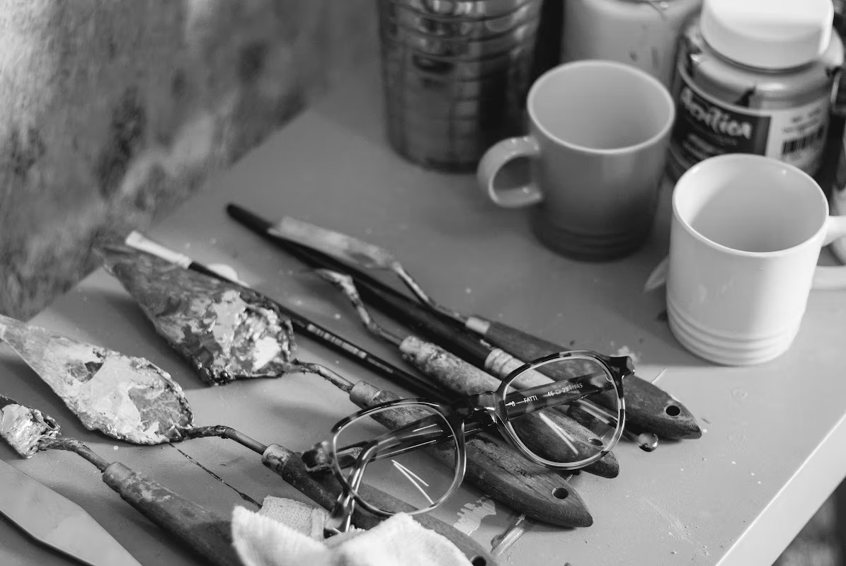 Ferramentas do ateliê de PH Monteiro — espátulas, pincéis, tintas acrílicas e óculos